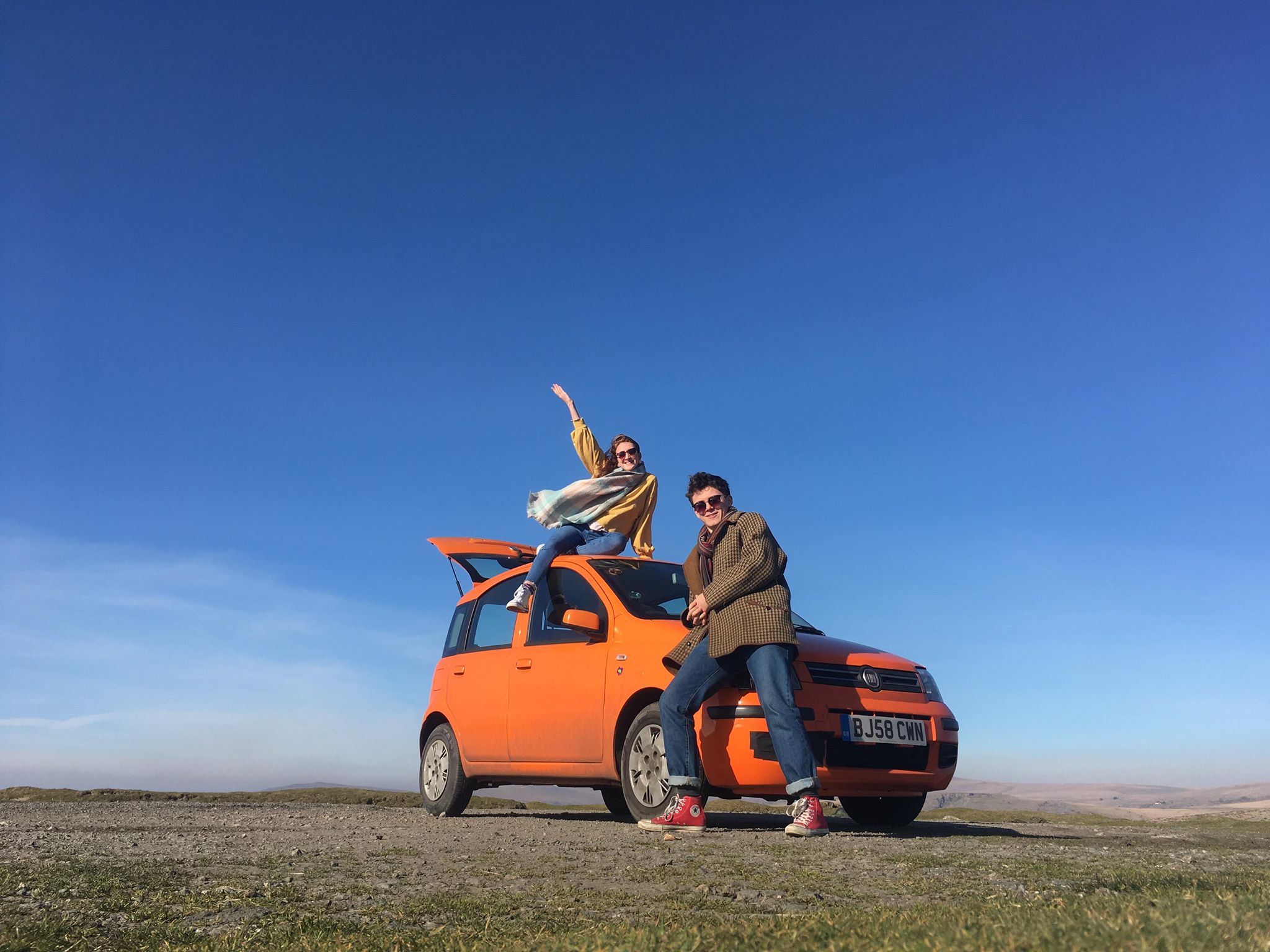 Lizzy and Matt posing on an orange car against a backdrop of blue skies and green grass.