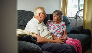 A couple in their fifties sit on a comfortable sofa, they are holding hands and talking.