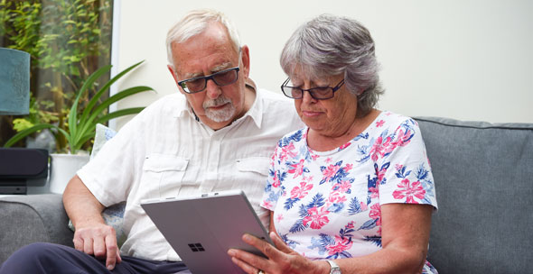 An older couple sit close together on a sofa using a tablet, they look comfortable and deep in thought.