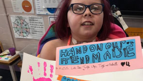 Maisie, a little girl diagnosed with a brain tumour, poses with a piece of art created for her by her classmates. The piece of art is called "Hand on our friend Maisie" and features children's handprints in bright paint alongside lines of poetry.