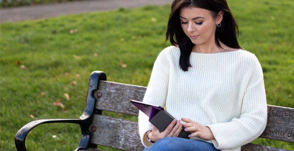 A young woman sits alone on a park bench using a tablet. She's dressed for warm weather and looks pensive.