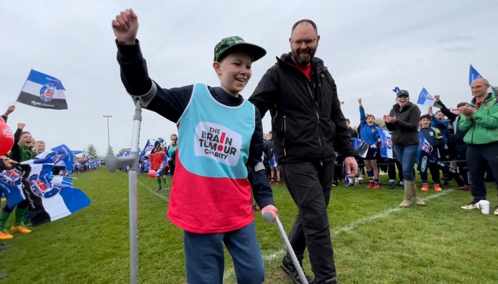 Will, an 11-year-old-boy, wearing a The Brain Tumour Charity, vest is celebrating completing his fundraising challenge. One fist is triumphantly raised in the air, while his other hand clutches a crutch to help him balance. His Dad, Neil, is looking on proudly as he walks beside him and a crowd can be seen cheering in the background.
